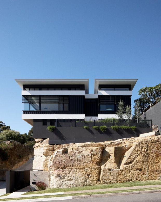 Exterior view of a contemporary multi-storey residence perched on a steep sandstone rock outcrop. The house features a strongly symmetrical composition, flat projecting roof planes, dark vertical cladding, white rendered bands, extensive glazing, balconies, and dark metal privacy screens. A driveway and garage entrance are visible below, with landscaped lawn and mature vegetation around the rocky site.
