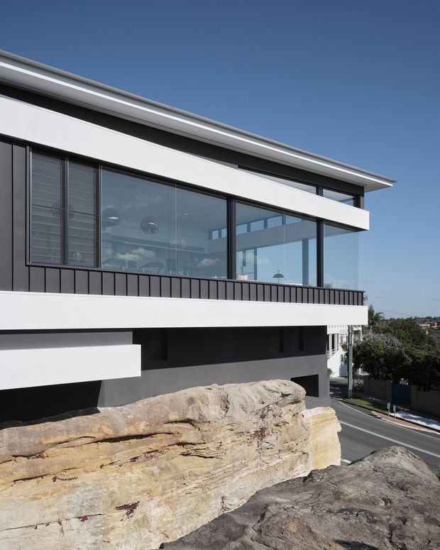 Contemporary multi-level residential building viewed from outside, with crisp white horizontal bands, dark charcoal cladding, extensive black-framed glazing, and a projecting flat roof. The structure is integrated into a rocky sloped site above a street, with a restrained modern facade and distant suburban surroundings.