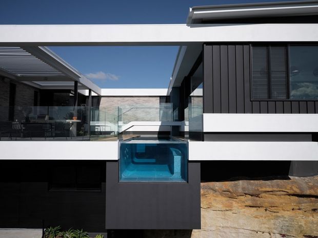 Exterior view of a contemporary multi-level residence built into a rocky sloping site. The design features crisp white horizontal bands, dark vertical cladding, extensive glazing, a glass balustrade, a covered outdoor terrace, and a projecting transparent blue swimming pool or spa element. A broad roof overhang and roller shutter are visible against a clear sky.