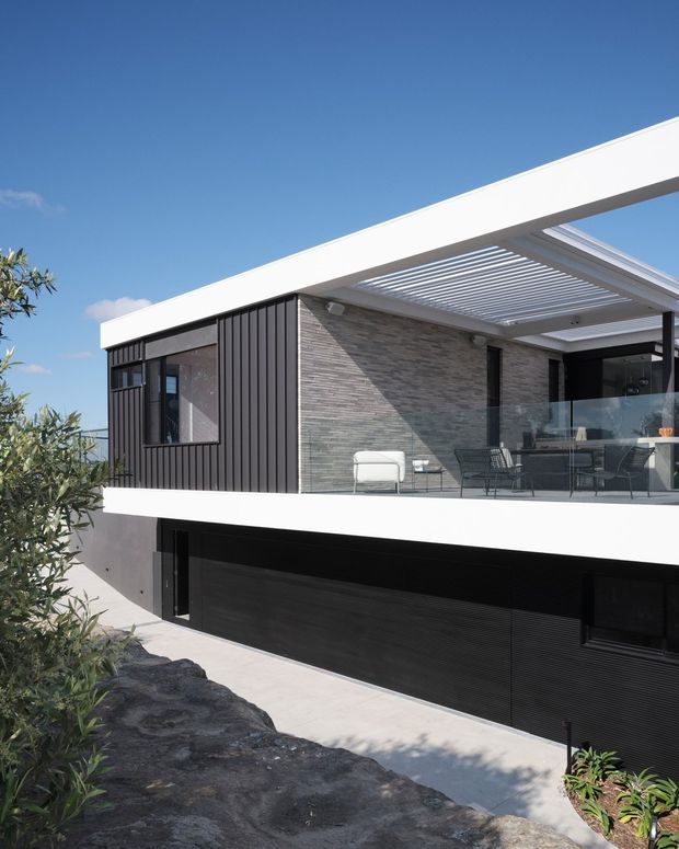 A modern two-level residential exterior with a flat roof and crisp white structural bands. The upper level combines dark vertical cladding with a grey stacked-stone feature wall, opening onto a covered terrace with a white pergola, glass balustrade, and outdoor seating. A large dark garage door occupies the lower level, with pale paving and landscaped planting in the foreground.