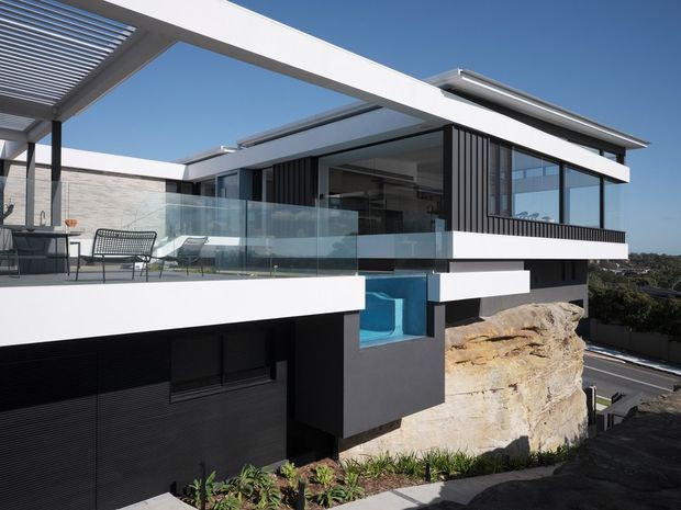 Exterior view of a contemporary multi-level residence built into a sloping rocky site. The house features crisp white horizontal volumes, dark textured wall cladding, extensive floor-to-ceiling glazing, black vertical screening, glass balustrades, rooftop or terrace pergola structures, and a vivid blue swimming pool visible beneath the upper terrace. Landscaping and exposed natural rock surround the lower level.