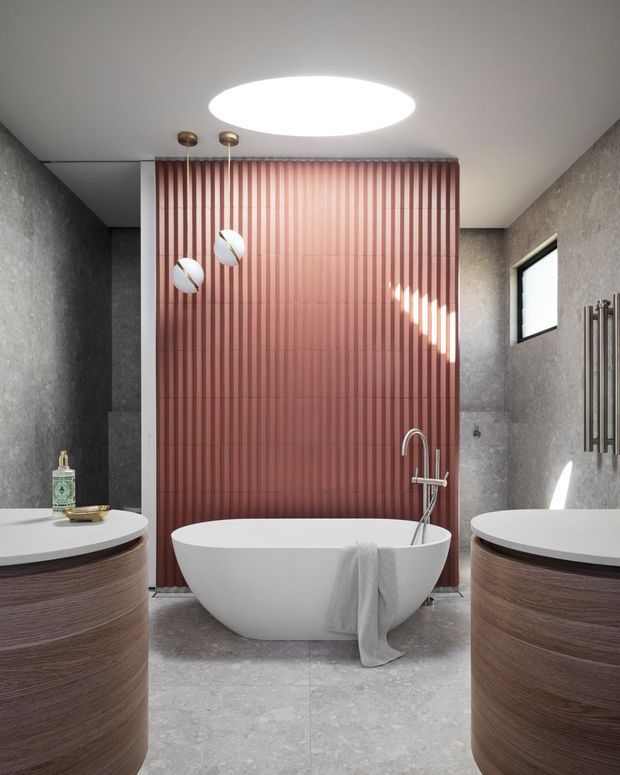 A contemporary bathroom featuring a freestanding white bathtub centered against a vertical ribbed pink-toned feature wall. Two rounded timber-veneered vanity units with white countertops frame the foreground, with a wall-mounted basin tap, dark textured wall finishes, large-format grey floor tiles, slim vertical towel radiators, a small horizontal window, and a large circular ceiling light. The composition is symmetrical and refined, with warm timber and blush accents balanced by cool grey surfaces.