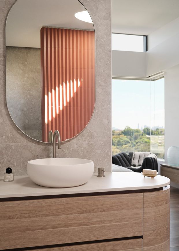 A refined contemporary bathroom vanity with a rounded white vessel basin, brushed metal wall-mounted tapware, and a large organically shaped mirror. The vanity features curved timber-look cabinetry and a pale stone or solid-surface benchtop. A textured neutral wall finish and warm terracotta vertical blinds add depth, while glazing opens the view toward a bright living area and landscaped outlook.