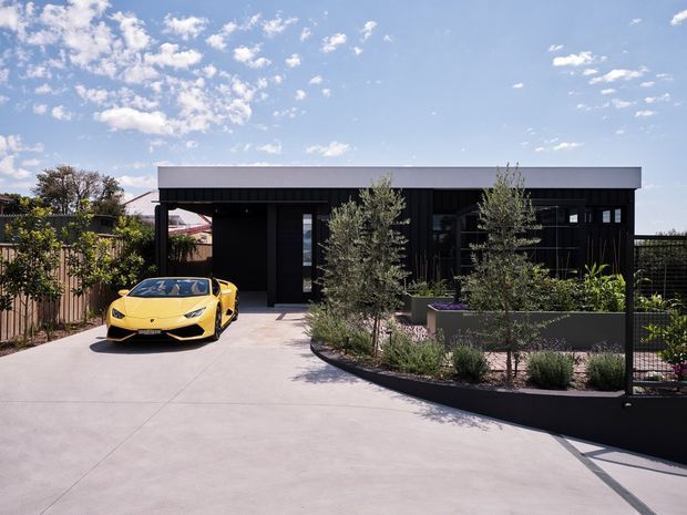 A single-storey modern black-clad residence with a flat roof, broad concrete driveway, dark-framed glazing and a landscaped front garden in raised planters. A yellow sports car is parked at the entrance, with fencing and neighbouring residential elements visible around the site under a bright blue sky.