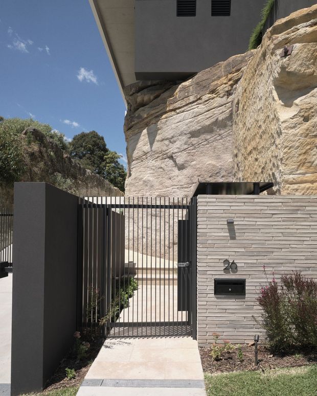 Street-facing entrance to a contemporary residence built directly against a prominent natural sandstone rock face. The image shows a narrow concrete walkway leading through a tall black metal slatted gate, with a dark rendered boundary wall and a pale horizontally layered masonry letterbox wall marked with the number 20. The building above has a projecting flat roof and dark ventilation grilles, while sparse landscaping borders the entry.