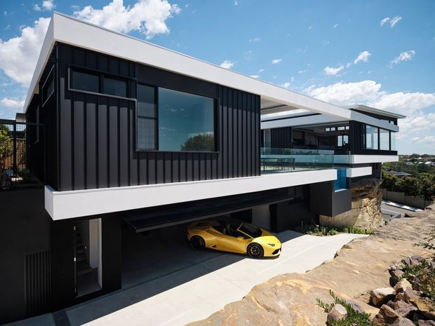Exterior view of a contemporary multi-level residence on a rocky sloped site. The home features bold cantilevered rectangular volumes, black vertical metal cladding, white fascia and trim, extensive dark-framed glazing, flat roofs, and an integrated undercroft garage with a car. Blue sky, distant suburban landscape, and exposed rock surround the building.
