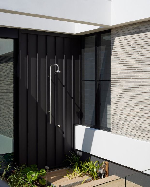 Modern residential entrance courtyard featuring a tall matte-black vertically ribbed door, a narrow chrome handle, black-framed glazing, light grey stacked-stone wall cladding, and crisp white rendered surrounds. A small planted entry bed and timber decking sit in the foreground, with strong sunlight and architectural shadows emphasizing the contemporary facade.