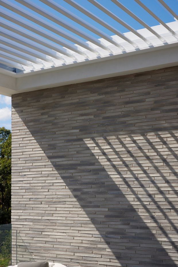 Close-up exterior architectural view of a contemporary masonry wall beneath a projecting pergola or canopy. The wall is built from horizontally laid narrow grey-brown bricks, while the canopy has white structural beams and translucent glazing or panels that cast strong linear shadows across the facade. Blue sky and a small amount of landscaping are visible at the edges.