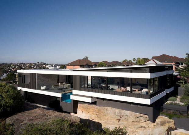A contemporary multi-level residence with a strongly horizontal, flat-roofed form, extensive floor-to-ceiling glazing, and dark vertical cladding accented by crisp white fascia bands. The house is elevated over a rocky, sloping site, with a terrace and outdoor seating visible on the upper level. Suburban houses appear in the background.