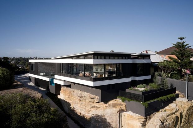 Exterior architectural photograph of a contemporary two-storey home built into a rocky, sloping site. The house has strong horizontal lines, dark cladding, broad white fascia bands, extensive glazing, a low-pitched roof, and an elevated terrace or balcony. Neighbouring suburban homes and landscaped planting are visible around the property.