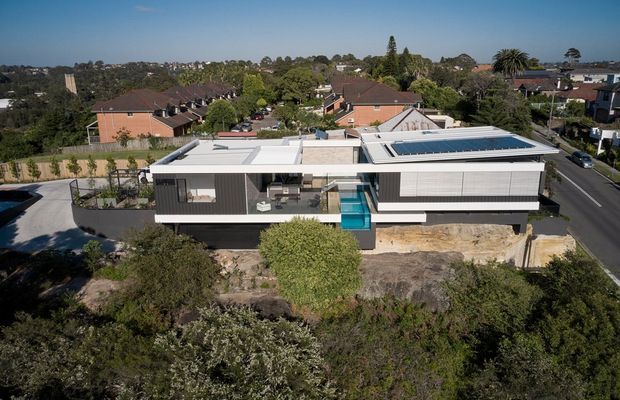 Aerial view of a large contemporary split-level residence on a suburban, sloping site. The house has a long low-profile form, flat roof sections, extensive horizontal glazing, dark and light cladding, a central turquoise swimming pool or water feature, and rooftop solar panels. Established vegetation and neighbouring suburban homes surround the property.