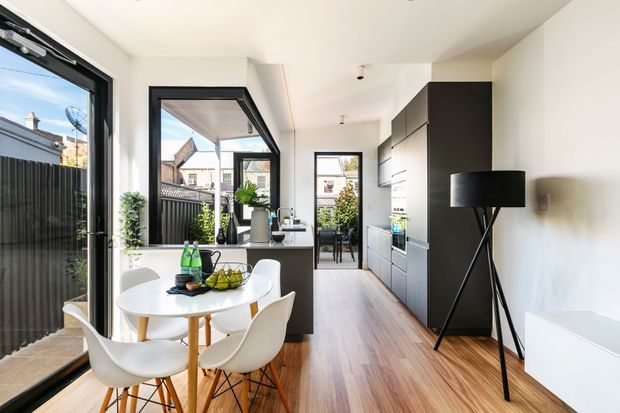 Bright, narrow contemporary open-plan kitchen and dining area with extensive black-framed glazing and a glass door opening to an outdoor area. The kitchen features a long run of dark built-in cabinetry, pale benchtops, integrated appliances, and a central island. A round timber dining table with white molded chairs occupies the foreground, while a black tripod floor lamp and white low cabinet sit along the wall. Timber flooring, white walls, and abundant daylight create an airy modern interior.