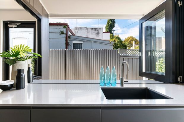 A contemporary kitchen work area with a large white stone benchtop, undermount black sink and brushed metal tap. Black-framed awning windows open onto a small urban courtyard, while dark flat-panel cabinetry runs below the bench. A potted plant and a few bottles provide minimal styling.