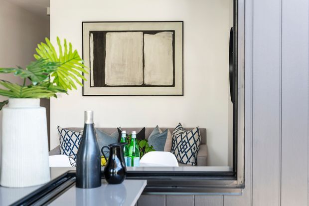 A close-up view of a refined contemporary living area seen through a large black-framed interior opening or reflective glazing. A pale sofa with patterned and neutral cushions sits against a light wall beneath a monochrome abstract artwork. In the foreground are a glossy pale counter or console surface, dark decorative bottles, green glass bottles, and a large white planter with tropical foliage. Vertical light-grey wall panelling or cabinetry appears at right, with black trim framing the opening.