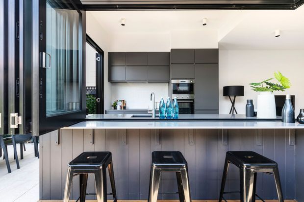 A contemporary kitchen viewed from an adjoining outdoor or glazed area, with a large dark island and three black metal stools in the foreground. The kitchen features matte dark-grey flat-panel cabinetry, a pale benchtop, integrated oven and microwave, stainless-steel sink tapware, recessed ceiling lights, and black-framed full-height glazing with an open hinged door. Minimal decorative objects and greenery add contrast.