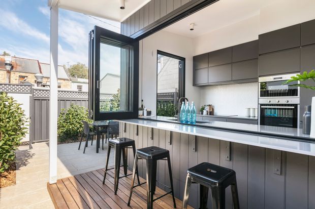 Bright contemporary kitchen opening onto a timber deck and paved courtyard through large black-framed folding or stacking doors. The kitchen features a long white stone island with black stools, dark grey flat-panel cabinetry, integrated appliances, a light tiled or painted splashback, and vertical dark wall panelling beneath the island. Outdoor fencing and planting are visible beyond the open glazing.