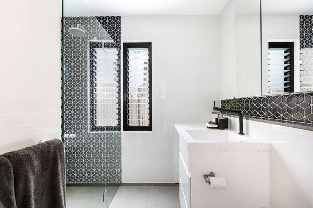 Bright contemporary bathroom with a frameless glass shower enclosure, black-and-white geometric patterned tile feature walls and splashback, white vanity with integrated basin, black tapware, mirrored wall, and plantation-shutter windows. A towel rail with a dark towel is visible in the foreground.