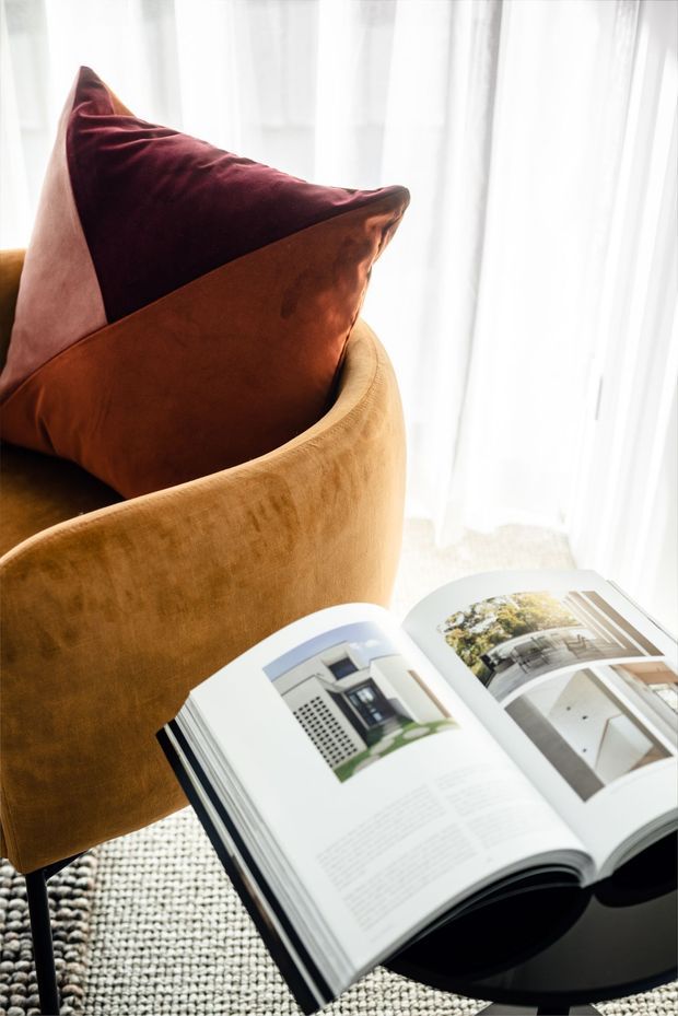 Close-up interior vignette of a plush mustard-tan upholstered armchair with burgundy and burnt-orange cushions beside a small round black side table. An open architectural or interior-design book rests on the table, with sheer white floor-to-ceiling curtains behind and a textured neutral rug beneath. The scene is warmly styled and softly lit.
