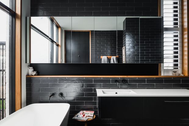 A modern bathroom featuring black subway-tile walls, a wall-mounted black vanity with an integrated white basin, a freestanding white bathtub, a large mirrored wall, timber-framed windows, and white horizontal blinds. The high-contrast monochrome palette is softened by warm timber accents and natural daylight.