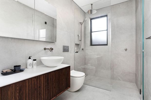 Contemporary bathroom with large-format light grey wall and floor tiles, a floating dark timber vanity with white benchtop, vessel basin, wall-mounted tapware, toilet, handheld and overhead shower, frameless glass shower screen, and black-framed window.