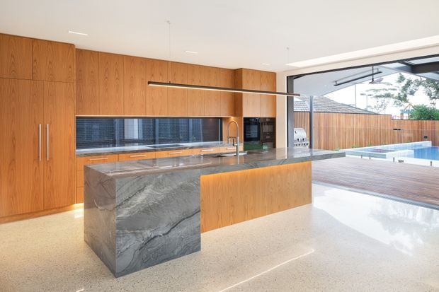 A spacious contemporary kitchen with full-height warm timber cabinetry, a dramatic veined stone island with waterfall end, integrated appliances, and a long linear pendant light. Polished terrazzo-style flooring extends toward large open glazing and a timber pool deck, with a swimming pool visible beyond.