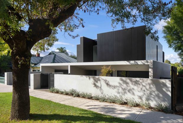 Street-facing view of a contemporary multi-level residence with a strong geometric composition. The home features smooth light rendered walls, a dark vertically ribbed upper volume, a flat roofline, recessed openings, a covered entry or carport-like opening, and a low front boundary wall. Mature street trees and a paved footpath frame the facade.