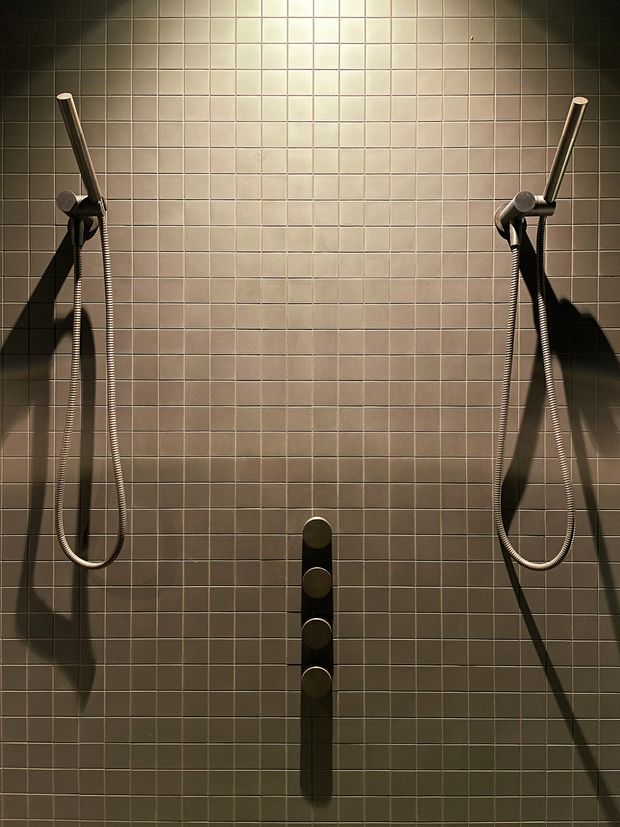 A symmetrical, close-up view of a tiled shower wall with two handheld shower fixtures and hoses mounted on either side, plus a central vertical row of dark circular controls. Dramatic directional lighting creates strong shadows across the neutral square-tile surface.