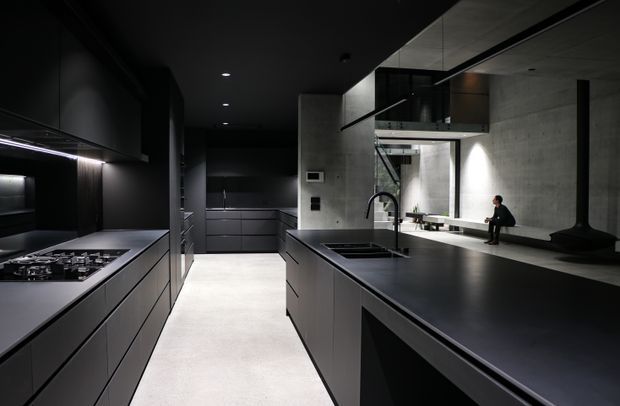 A dramatic, highly minimalist contemporary kitchen with dark matte cabinetry, expansive black benchtops, integrated appliances, and a large central island with sink. The space features polished concrete walls, a pale smooth floor, concealed linear lighting, and a glazed upper-level void or mezzanine visible beyond the kitchen. A sculptural black fireplace or flue appears along the rear wall, with a person seated on the deep rear ledge.