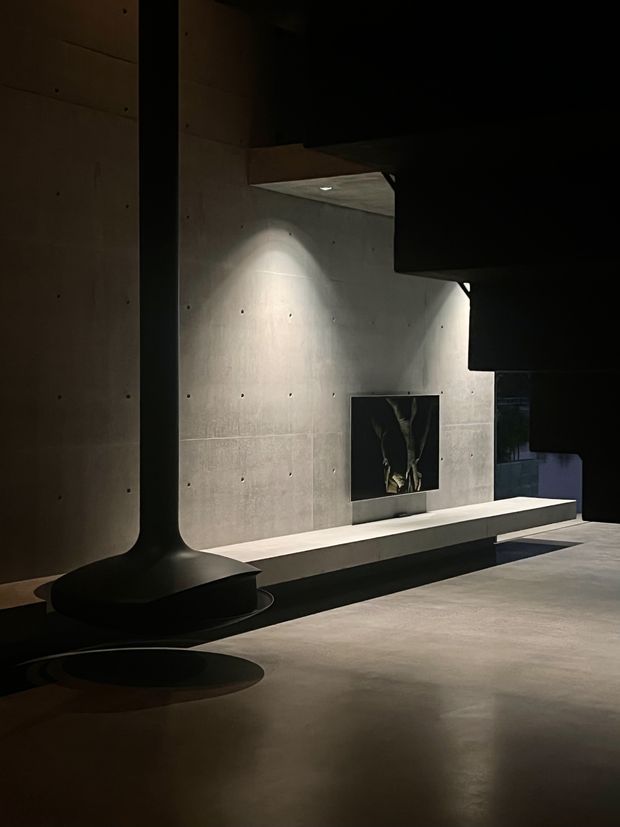 A dramatic, dimly lit contemporary interior with exposed smooth concrete walls, floor, and sculptural architectural forms. A long built-in concrete bench runs along the wall beneath a dark framed artwork, while recessed lighting creates focused pools of illumination. The space has a gallery-like, minimalist atmosphere with strong geometric shadows and black structural elements.