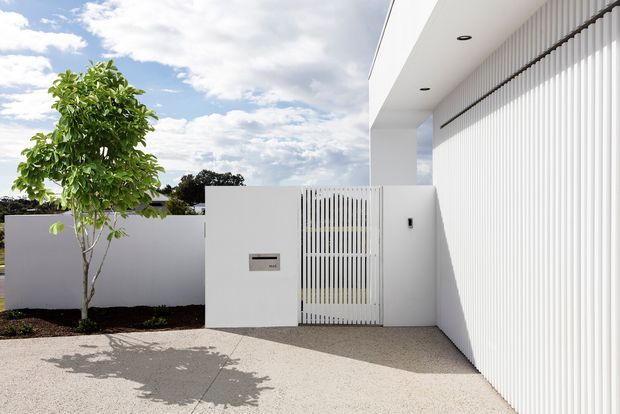 Bright, minimalist modern residential entrance viewed from the exterior. A white rendered boundary wall and pedestrian gate lead into a covered entry beneath a projecting flat roof. The facade features extensive vertical white slatted screening, recessed ceiling downlights, a letterbox, and a pale textured paved forecourt with a small planted tree. The composition is clean and highly geometric, with strong white surfaces and crisp shadows.