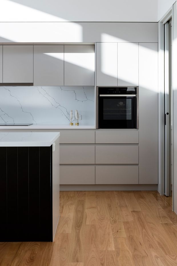 A bright contemporary kitchen with streamlined light-grey full-height cabinetry, integrated black wall oven, white veined stone splashback and benchtops, and a contrasting black island base. Warm timber flooring and strong sunlight entering from the right create crisp geometric shadows across the minimal joinery.