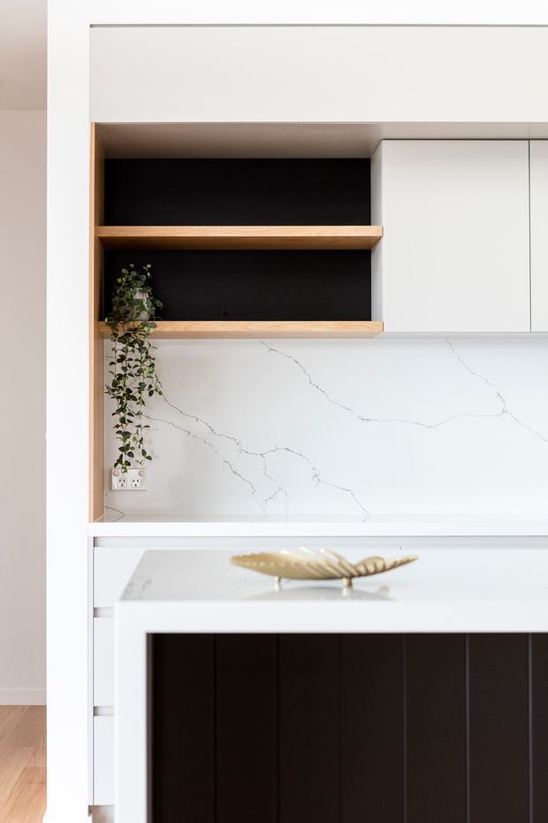 Close-up interior view of a contemporary kitchen featuring white cabinetry, open timber shelves with black backing, and a white veined stone splashback and benchtop. A dark vertical-panel island front and a small trailing plant add contrast to the restrained monochrome and timber palette.