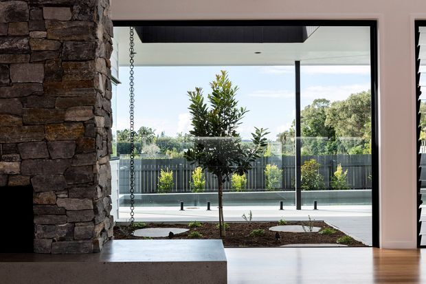 View from a contemporary interior through full-height glazing toward a covered outdoor terrace and landscaped courtyard. A rugged stacked-stone feature wall and hearth occupy the left side, while the interior has smooth pale walls, a dark ceiling soffit, and a polished light floor. Outside, a small tree, planted garden beds, glass balustrade, and dark vertical fencing are visible.