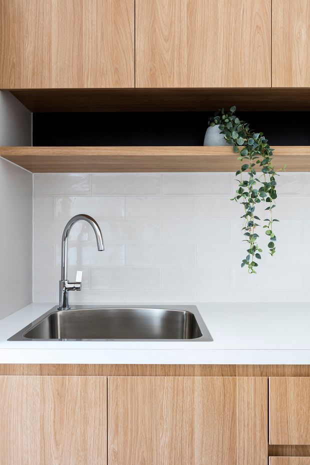 Close-up view of a compact contemporary kitchen centered on a stainless-steel undermount sink and curved chrome mixer tap. The kitchen features light timber-grain cabinetry above and below, a white smooth benchtop, glossy white subway-tile splashback, and an open dark timber shelf with a trailing potted plant.