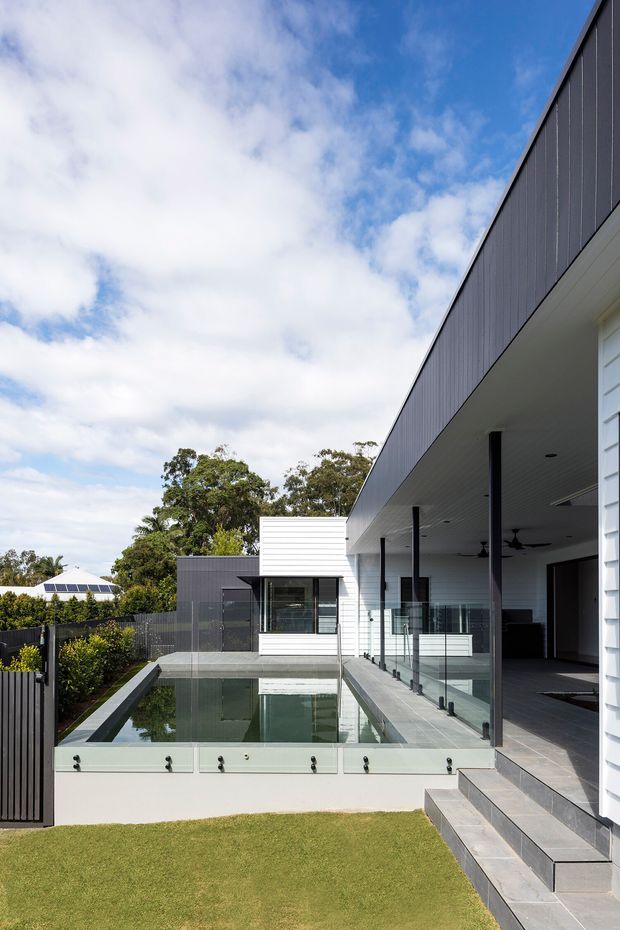 Exterior architectural photograph of a contemporary multi-level residence. The view shows a dark vertically clad upper facade, white horizontal weatherboard walls, deep roof overhang, slim black columns, glass balustrades, a covered outdoor living area, and a long rectangular reflecting or swimming pool beside tiled steps. A manicured lawn, fencing, neighbouring roof and trees are visible in the background beneath a partly cloudy sky.