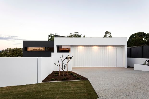 Exterior view of a modern single-storey residence at dusk, featuring a crisp white rendered facade, dark contrasting cladding, a broad double garage, minimalist landscaping, a pale gravel driveway, and clean horizontal lines.