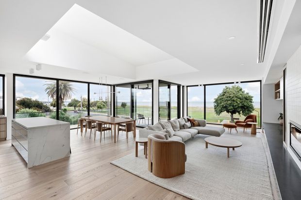 Bright open-plan contemporary interior combining a kitchen, dining area and living room, with expansive black-framed floor-to-ceiling glazing overlooking a landscaped waterfront or coastal setting. The space has pale timber flooring, a white marble-look kitchen island, a large light sectional sofa, curved timber lounge chairs, a dining table, and a large skylight or roof opening. The restrained palette and abundant daylight create an airy, refined atmosphere.