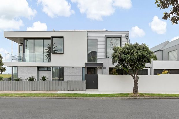 Street-facing view of a contemporary two-storey residence with a crisp white rendered facade, extensive glazing, a projecting upper-level balcony with clear glass balustrade, and horizontal light-grey cladding. A dark entry door, privacy wall, slim metal fencing, and integrated garage are visible, with a neighbouring pitched-roof home at right.