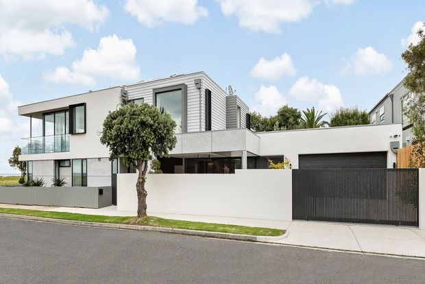 Street-facing view of a large contemporary multi-level residence with a predominantly white rendered facade, strong horizontal lines, extensive dark-framed glazing, a glass balcony balustrade, and a projecting upper floor. The home includes a section clad in light horizontal weatherboards, a dark panelled garage door, and a tall dark metal driveway gate. A neatly landscaped verge and mature trees sit in front of the property.