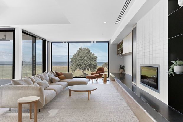 Bright, refined living room with a large cream sectional sofa, round timber coffee tables, and sculptural lounge chairs arranged around a contemporary fireplace. Full-height glazing opens the room visually toward a coastal ocean view, while a built-in shelving and fireplace wall combines pale tiled surfaces with dark stone trim. The space has a calm, airy, minimalist aesthetic with abundant natural light.