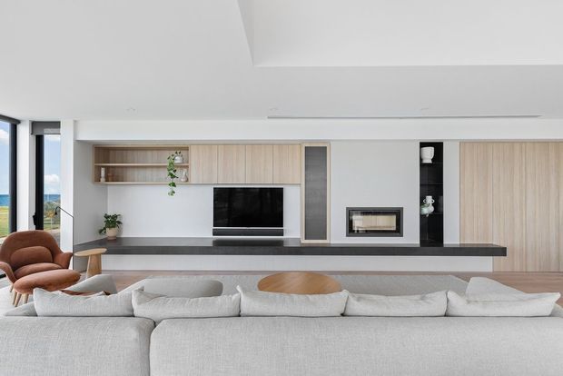 Contemporary living room viewed from behind a large light-grey sectional sofa, featuring a full-width built-in timber and white media wall with open shelving, television, fireplace, dark stone ledge, and concealed storage. Floor-to-ceiling glazing at left brings in a bright coastal-looking view, while a sculptural brown lounge chair and simple timber side table add warmth.