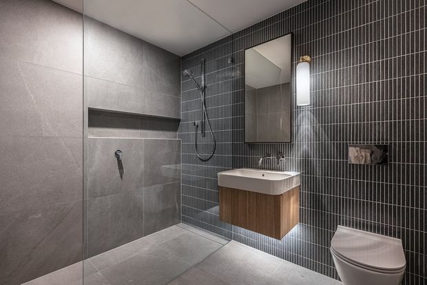 Contemporary bathroom with a walk-in glass shower, large-format grey wall and floor tiles, a dark vertically tiled feature wall, floating timber vanity with white basin, wall mirror, vertical wall light, and concealed-style toilet.