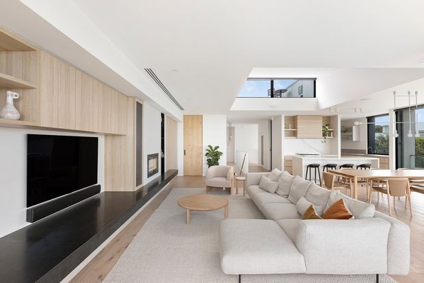 Bright, open-plan contemporary living area with a large pale sectional sofa facing a black built-in media wall. Light timber wall panelling and joinery extend toward a dining area and white kitchen with an island and stools. A skylight or double-height opening brings natural light into the room, while large windows provide additional daylight. The palette is predominantly white, cream, pale timber, and black accents.