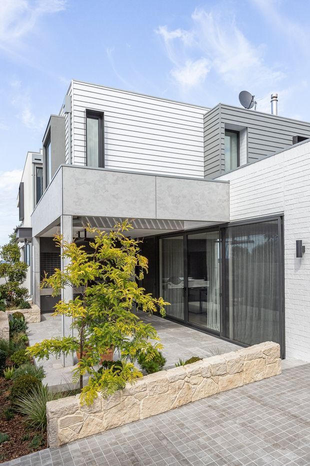 A contemporary multi-level residential exterior with crisp white horizontal cladding, white brickwork, dark-framed floor-to-ceiling glazing, and a projecting concrete balcony or canopy. The property features a paved courtyard and walkway, low sandstone retaining walls, and landscaped planting. The bright facade is photographed under a blue sky.