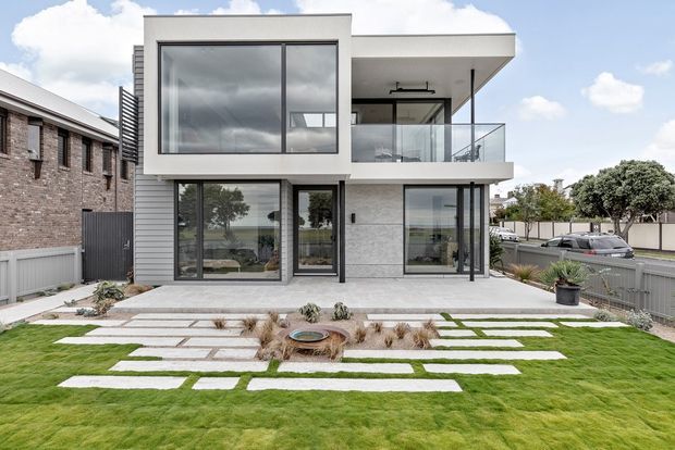 Contemporary two-storey residential house viewed from the front, featuring a crisp white rendered facade, grey horizontal cladding, extensive dark-framed glazing, a projecting upper-level volume, and a glass-balustraded balcony. The front landscape uses a geometric arrangement of large pale paving slabs, lawn strips, gravel, and low ornamental planting. A neighbouring brick building and street are visible at the sides.