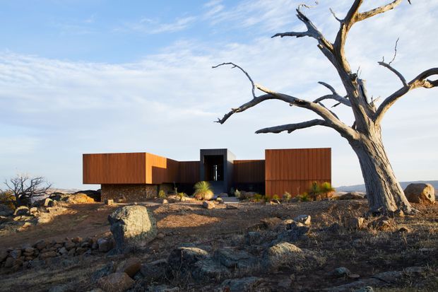 A low, angular modern house set in a stark rocky desert landscape. The building has rust-coloured vertical cladding, dark recessed central glazing, and stone-clad lower walls, with a dramatic dead tree dominating the foreground.