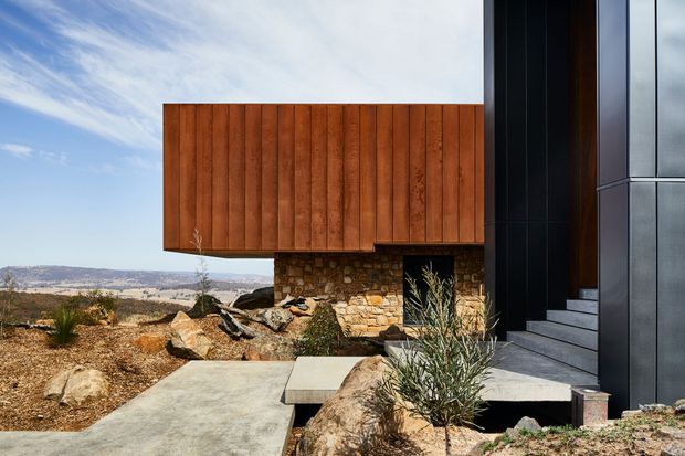 Contemporary angular house exterior set in a dry, rocky landscape. The facade combines warm vertical timber cladding, rough natural stone masonry, and dark metal panels around a recessed entry with broad concrete steps. A concrete path crosses the xeriscaped foreground, with distant open hills visible beyond the building.
