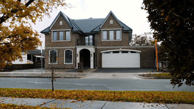 Street-facing view of a substantial two-storey brick residence with a symmetrical gabled roof, dark roof tiles, arched entry porch, tall multi-pane windows, and an integrated double garage. Autumn trees and fallen leaves frame the suburban frontage.