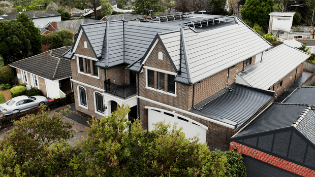 Aerial view of a substantial multi-storey suburban brick residence with multiple steep gabled roof sections, dark tiled and light metal roofing, white trim, balconies, and an attached double garage. Solar panels are visible on the main roof, with surrounding mature vegetation and neighbouring homes.