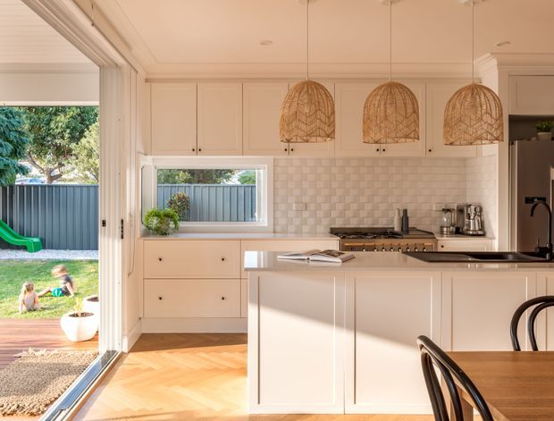 A bright, warm kitchen with cream-painted cabinetry, a white tiled backsplash, timber herringbone flooring, and a large central island with a dark benchtop. Three woven pendant lights hang above the island. The kitchen opens directly to a grassy outdoor area through large glazed doors, with a dining table and chairs visible in the foreground.